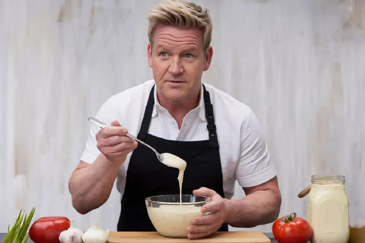 A renowned chef in a black apron holding a spoon and mixing creamy sauce in a bowl, surrounded by fresh ingredients like garlic and tomatoes.